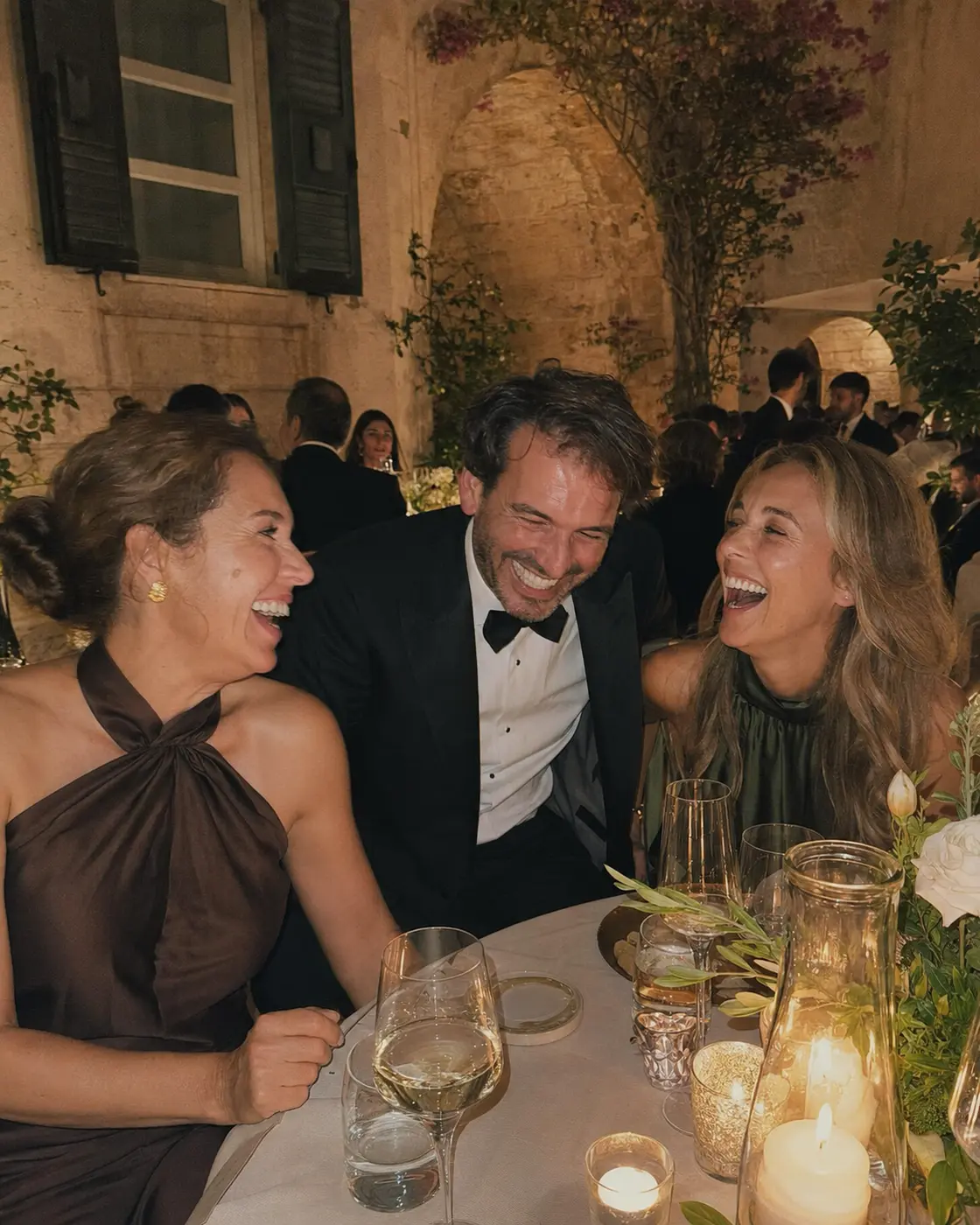 Three adult friends laughing over dinner in a stone courtyard